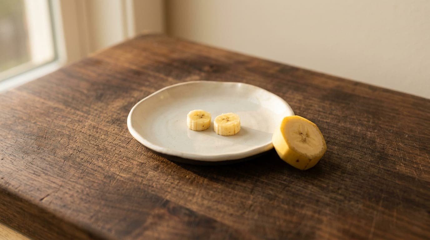 Two pea-sized pieces of fresh banana on a small white ceramic dish, beside a banana slice for scale